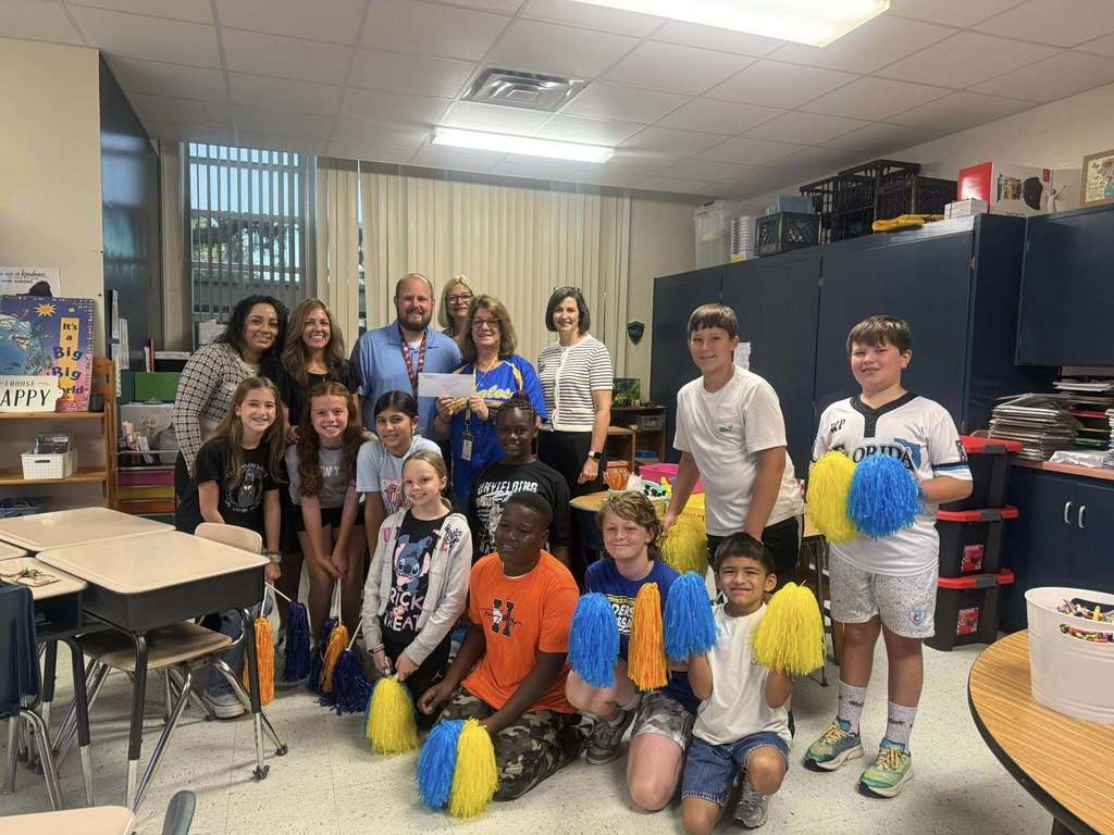 In a classroom, a group of approximately 14 students and several adults, with some holding blue and yellow pom-poms and a large check, stand around student desks.