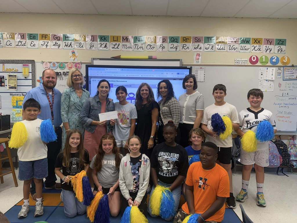 A group of students holding blue and yellow pom-poms kneel or stand with several adults in front of an interactive whiteboard displaying educational content in a classroom.