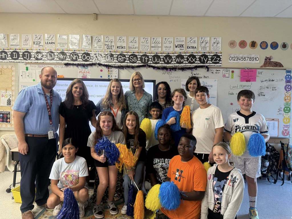 A group of students holding blue and yellow pom-poms kneel or stand with several adults in front of an interactive whiteboard displaying educational content in a classroom.