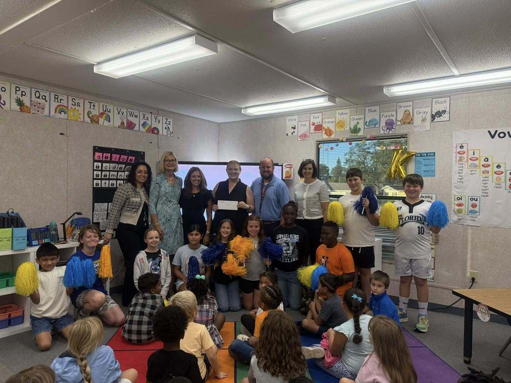 A group of students holding blue and yellow pom-poms kneel or stand with several adults in front of an interactive whiteboard displaying educational content in a classroom.