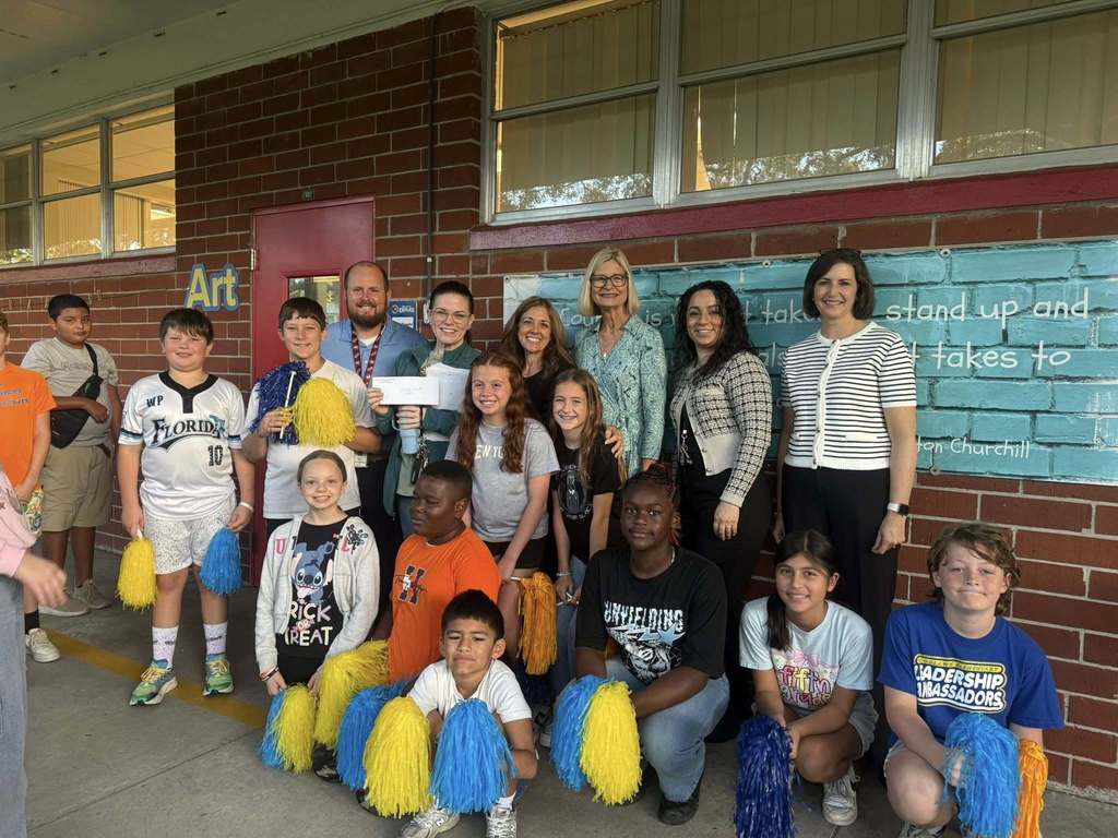 A group of approximately 16 students and several adults, some holding blue and yellow pom-poms and a large check, pose for a photo outside a school building with a brick wall and a teal-painted mural.