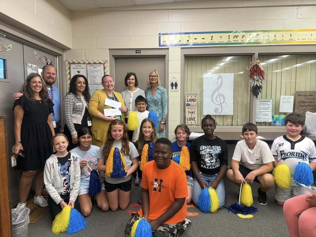 A group of students holding blue and yellow pom-poms kneel or stand alongside several adults in a classroom setting with a closed door, large windows, and a music-themed decoration.