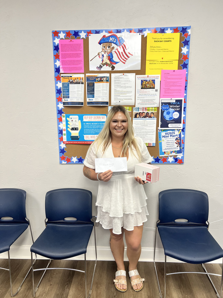 A smiling woman stands indoors in front of a colorful bulletin board decorated with red, white, and blue stars. She is holding an envelope and a small prize box. The bulletin board behind her displays school-related flyers and a cartoon mascot holding an American flag. Three blue chairs are lined up along the wall.