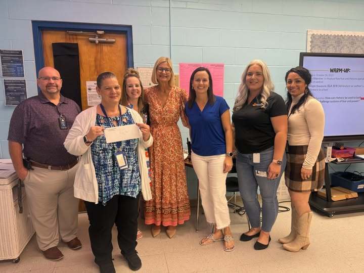  This image shows six women and one man standing in a classroom. A woman in the center, wearing a white cardigan over a blue top, holds up a white envelope, possibly a check or award. They are posing in front of a colorful bulletin board and a screen displaying a "Warm-Up" lesson plan.