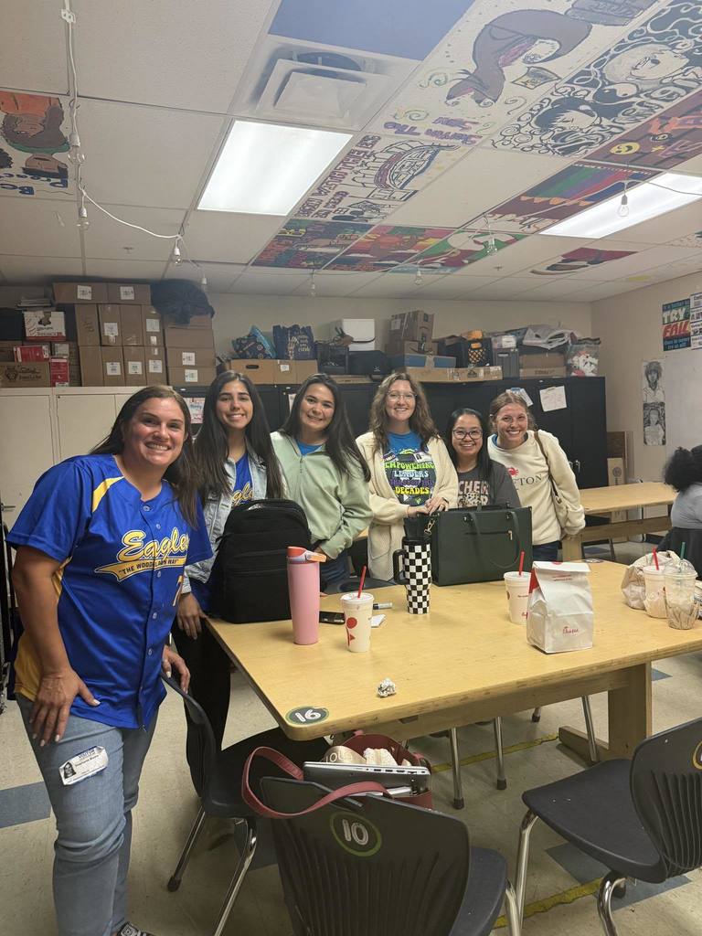 Six individuals, including one adult in a blue and yellow "Eagles" jersey, stand smiling behind a table in a classroom. The classroom features an artfully decorated ceiling, storage shelves stacked with boxes, and several chairs. Cups and a Chick-fil-A bag are on the table.