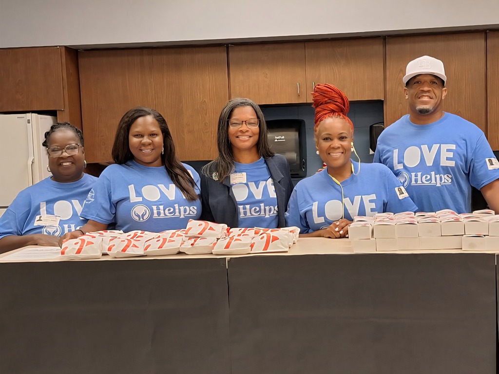 Five people are standing behind a counter in an indoor space. Four are wearing light blue "LOVE Helps" t-shirts. They are posing with stacked boxes and bags of food on the counter. The background is a kitchen area with wooden cabinets.