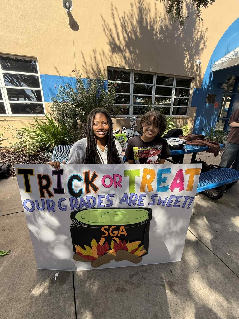 Two smiling students, one with long braids and one with curly hair, hold a large, colorful sign outdoors in front of a school building. The sign reads: "TRICK OR TREAT! OUR GRADES ARE SWEET!" Below the text is a drawing of a cauldron over a fire with "SGA" (Student Government Association) written on it.