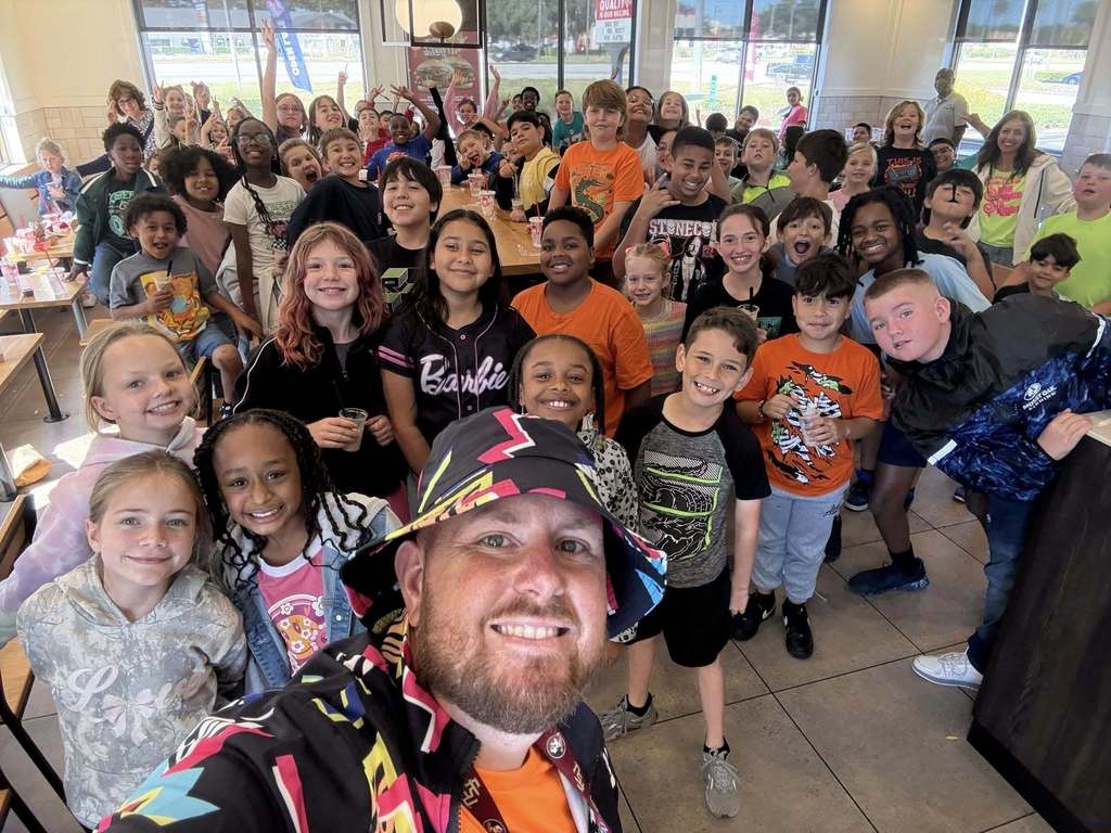 A bearded man wearing a colorful, patterned bucket hat and jacket takes a selfie with a large, diverse group of approximately 80 smiling children inside a brightly lit restaurant. The children are excited, waving, and looking at the camera. Tables and windows are visible in the background.