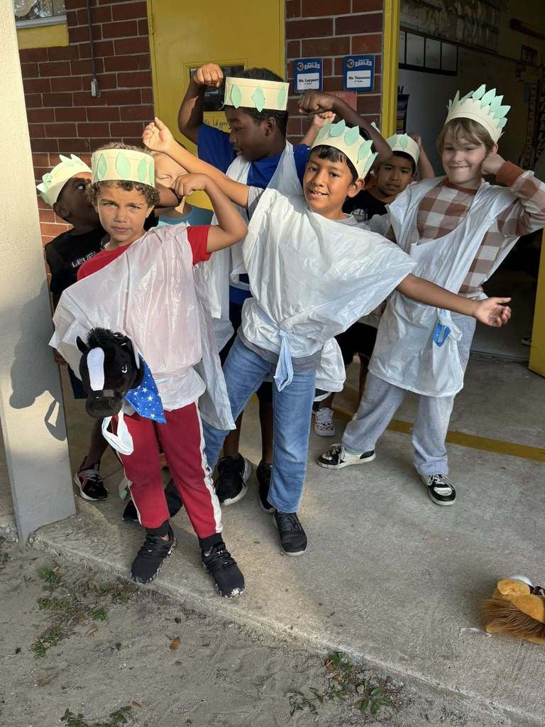 A group of approximately seven young boys is dressed in costumes, possibly for a history or culture event, standing outside a building. Several boys wear crowns resembling the Statue of Liberty's and white poncho-like coverings. They are smiling and posing, with some flexing their muscles. One boy holds a horse toy.