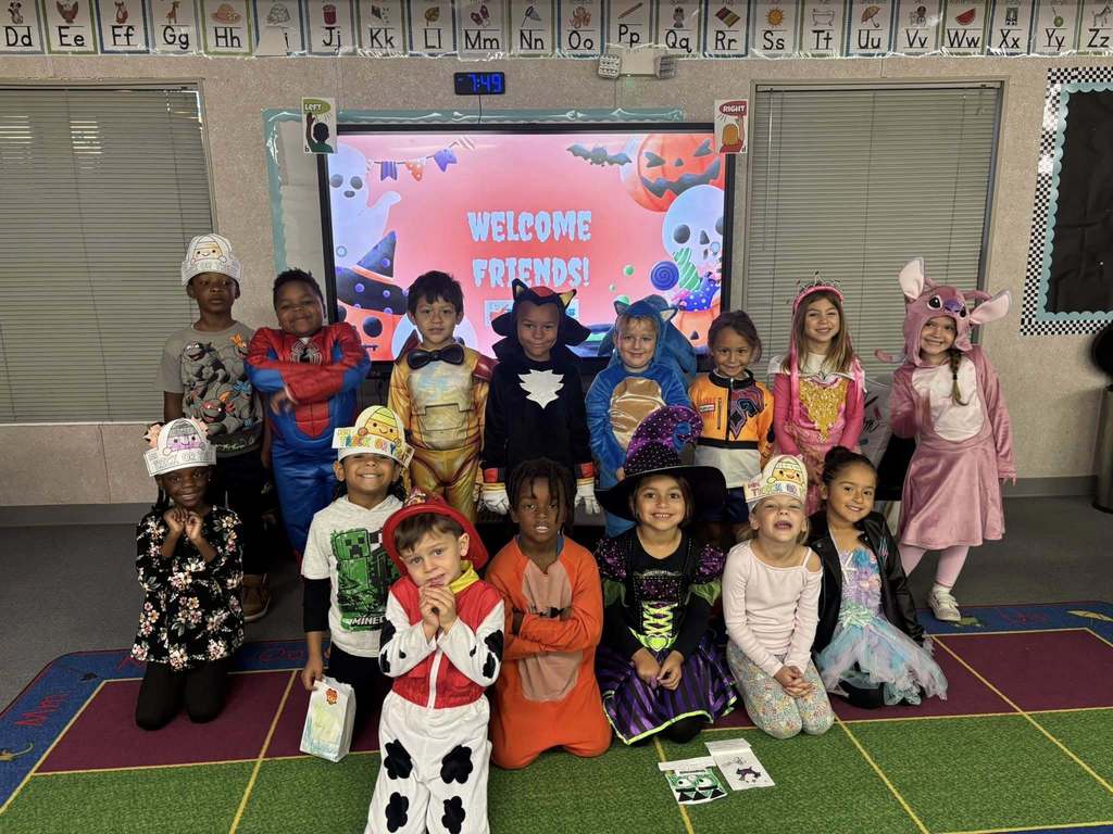 A group of approximately 14 young, diverse elementary school students is standing and kneeling in a classroom, all wearing Halloween costumes. They are posing in front of a large screen displaying a festive "WELCOME FRIENDS!" message with Halloween graphics.