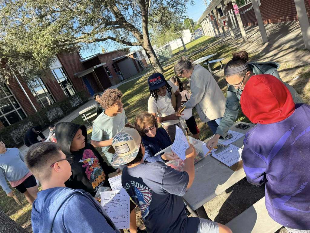A group of seven students and two adults are gathered around a gray picnic table outdoors on a sunny day. They are engaged in an activity, looking at papers, and interacting with the adults who appear to be helping them or directing the task. The background shows a school building and trees.