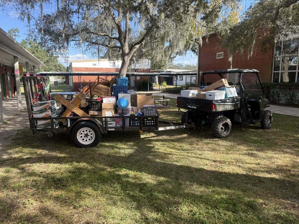 A Polaris Ranger utility vehicle is parked on a grassy area, towing a small trailer loaded with various items, including boxes, crates, and a globe, suggesting a move or delivery. The scene is outdoors on a sunny day near a brick school building with covered walkways and an oak tree with Spanish moss.