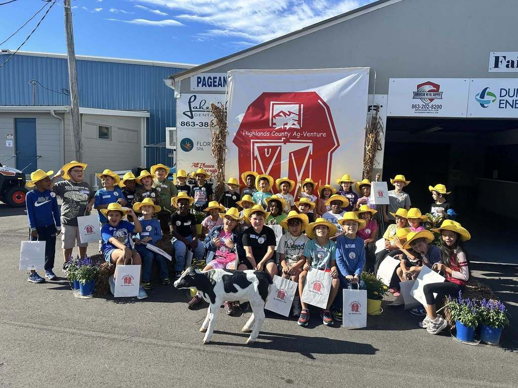 A group of approximately thirty elementary-aged students and a small calf pose outdoors. The children are all wearing matching yellow cowboy hats. Behind them is a large banner with a red barn silhouette and the text "Highlands County Ag-Venture."