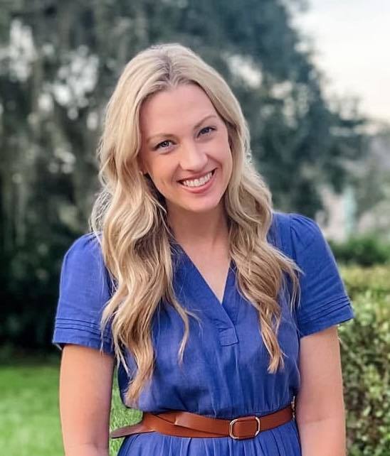 A headshot of a smiling, blonde woman with long, wavy hair, standing outdoors against a soft-focus background of trees and greenery. She is wearing a short-sleeved blue dress with a V-neck and a brown belt.