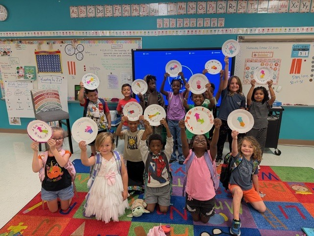 MES primary class students holding up paper plate clocks they have made to help with telling time.