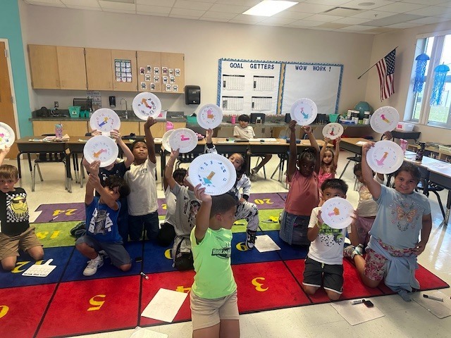 MES primary class students holding up paper plate clocks they have made to help with telling time.