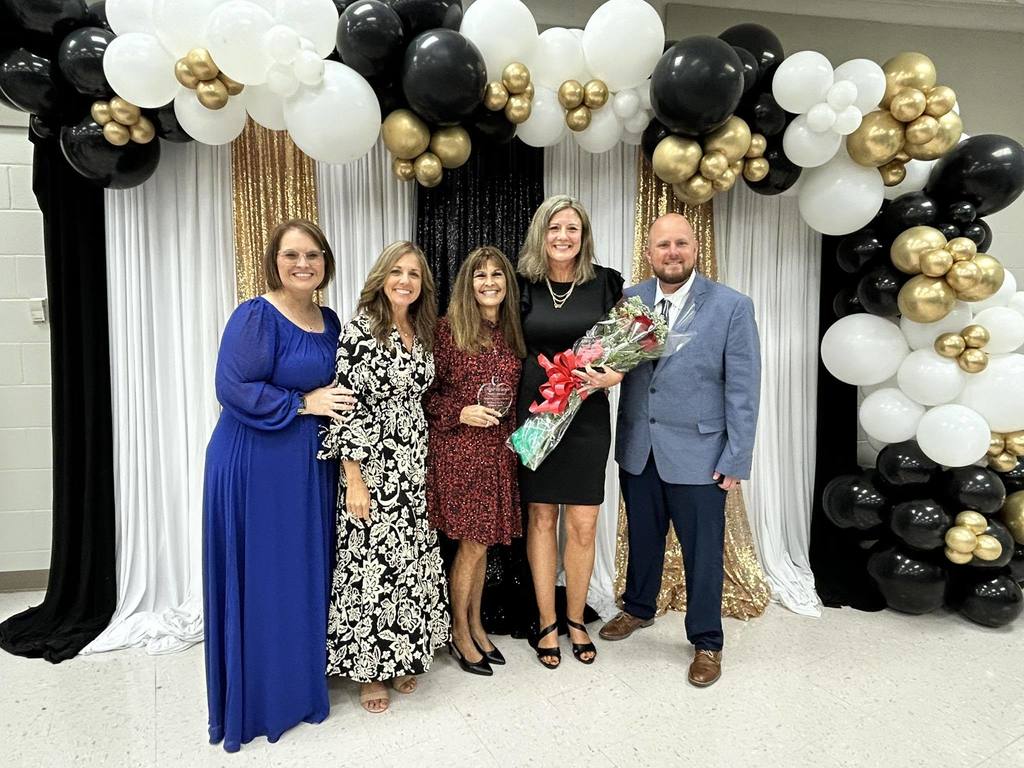 A photograph of five adults, three women and two men, standing in front of a celebratory backdrop. The backdrop features black and white draping, gold sequin fabric, and an arch of black, white, and gold balloons. The woman in the center holds a small award, and the woman next to her holds a bouquet of red roses.