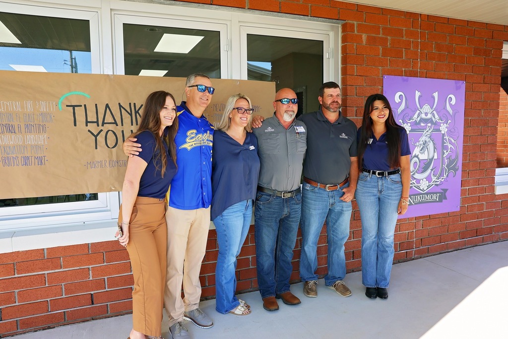 A photo of six people—three women and three men—standing outside against a brick wall. They are gathered in front of two banners. The large brown banner says "THANK YOU" and lists various names. A smaller purple banner features a school mascot. All six people are smiling and appear to be posing for a group photo.