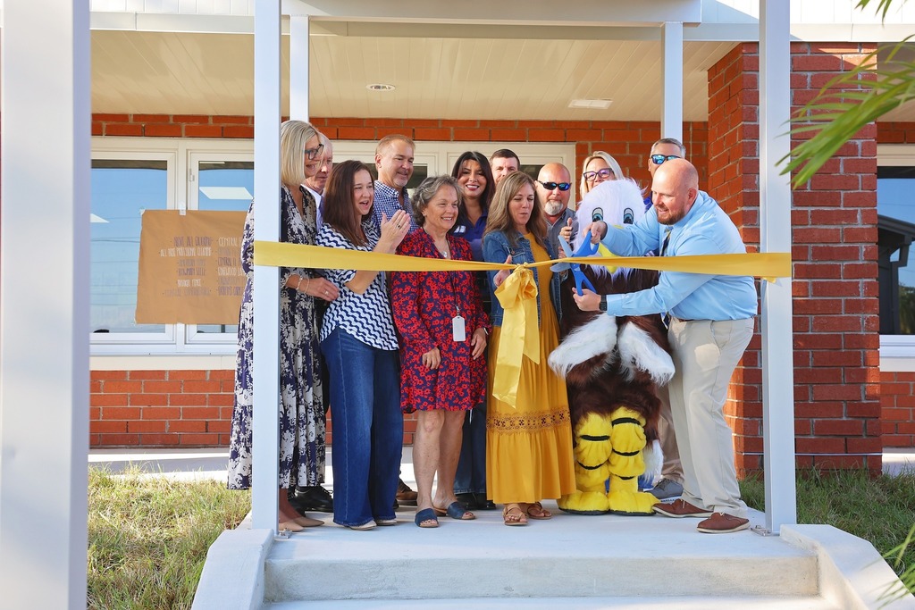 A ribbon-cutting ceremony with a group of approximately ten adults and a large eagle mascot. They are gathered on a porch in front of a brick building. A man in a blue shirt and khaki pants, with the help of the mascot, is cutting a yellow ribbon held by the group. A banner is visible through a window behind them.
