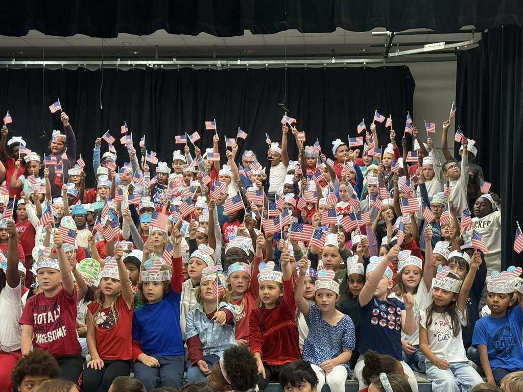 A large group of elementary school children, possibly on a stage or in an auditorium, sitting or standing. Most children are wearing patriotic paper crowns and enthusiastically holding small American flags high above their heads in celebration, against a background of black curtains.