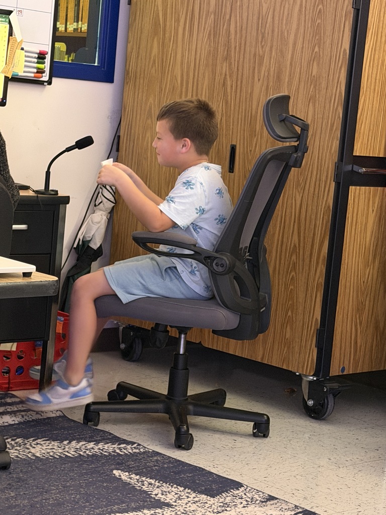 A boy with light brown hair, wearing a white patterned shirt and light blue shorts, sits in an office chair. He holds a white card while looking down, next to a desk with a microphone.