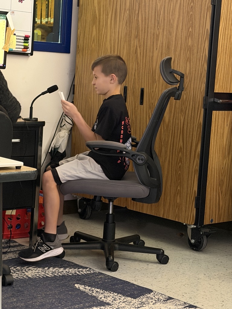 A boy wearing a black t-shirt and light gray shorts sits in a gray office chair. He holds a small white card in front of him, next to a black desk with a small microphone.