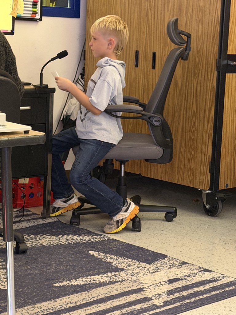 A boy with blonde hair, wearing a light gray hoodie and jeans, sits on the edge of a gray office chair. He is holding a small white card, positioned near a desk with a microphone.