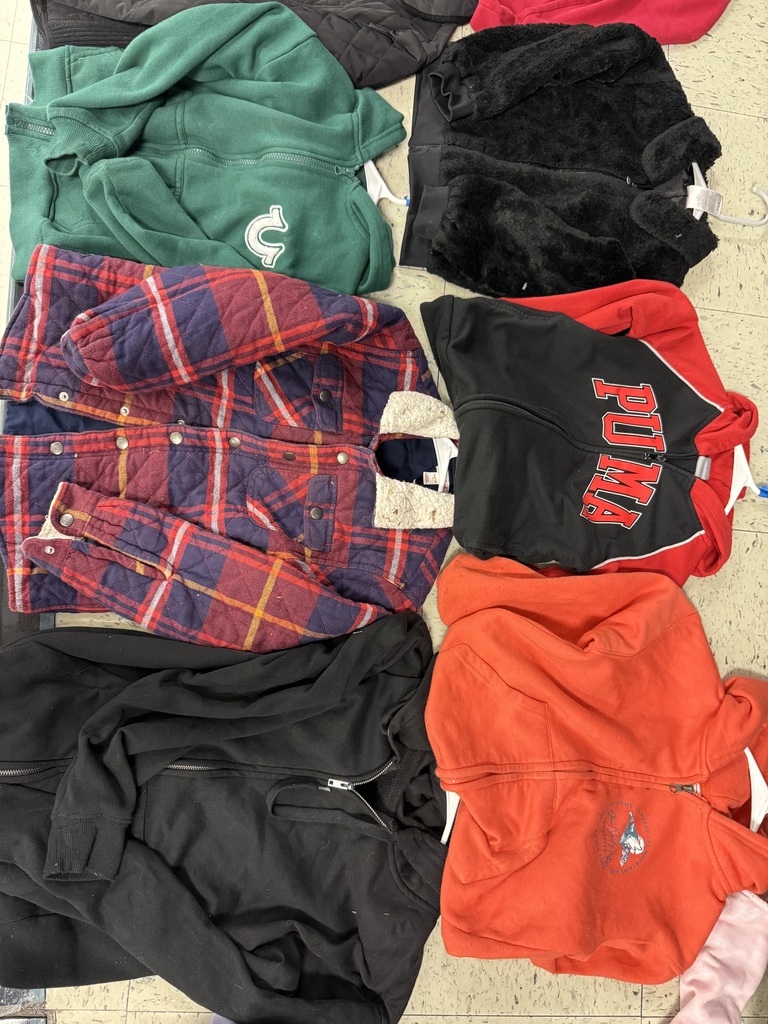 A pile of outerwear on a tiled floor, including a green fleece, a red and blue quilted plaid flannel jacket with a sherpa collar, a black zipper hoodie, an orange zipper hoodie, a black fuzzy jacket, and a red and black PUMA zipper jacket.