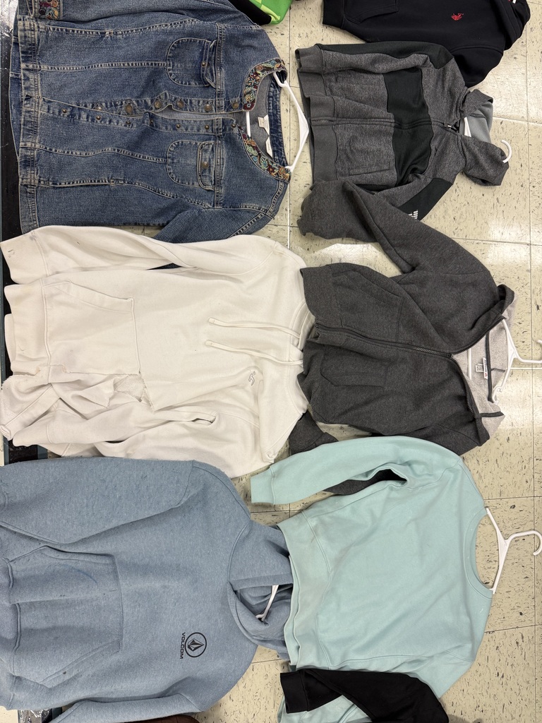 A pile of hoodies and jackets: a denim jacket with embroidered trim, a faded light blue hoodie, a cream hoodie, a dark gray full-zip hoodie, and a light mint green crewneck sweater.