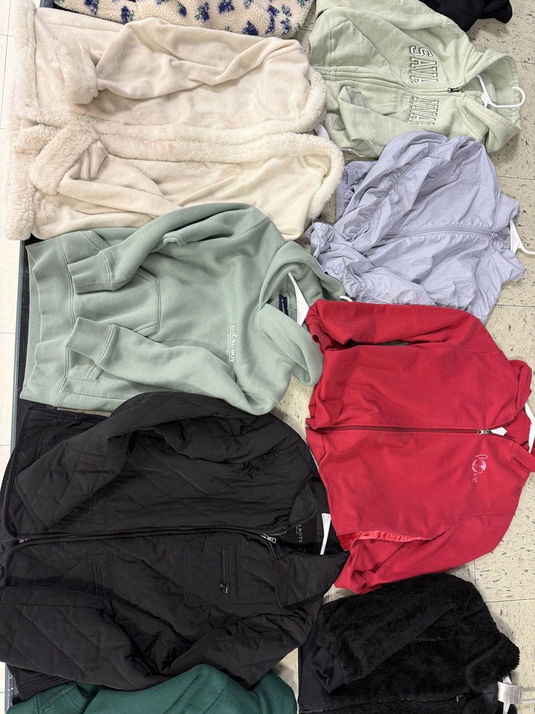 An overhead view of jackets and sweaters including a light tan sherpa coat, a sage green sweatshirt, a black quilted jacket, a red fleece, a black fuzzy jacket, and a crumpled lavender jacket.