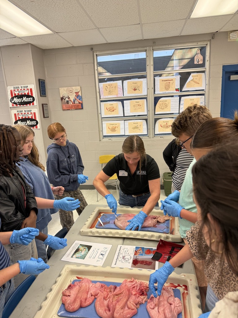 A female instructor wearing a black shirt and blue gloves demonstrates dissection techniques on a specimen to a group of female students gathered around a table.