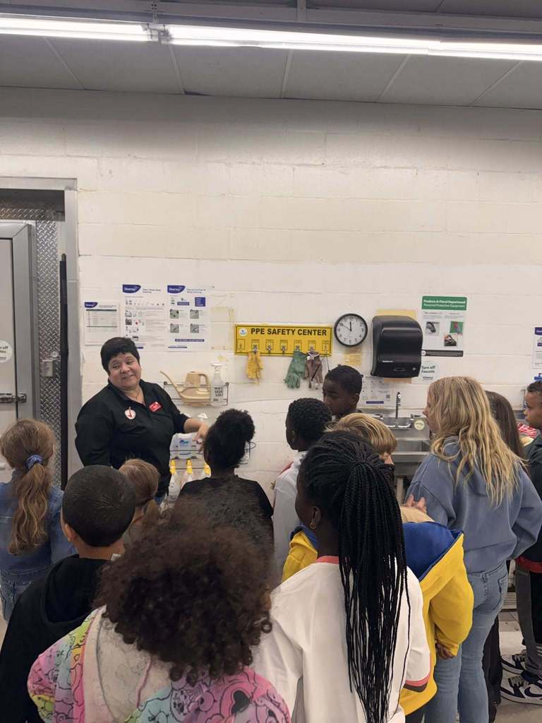 An adult woman in a black shirt is speaking to a group of school children in an industrial-looking room, likely a back kitchen or food prep area, next to a sink and an "PPE Safety Center" sign.