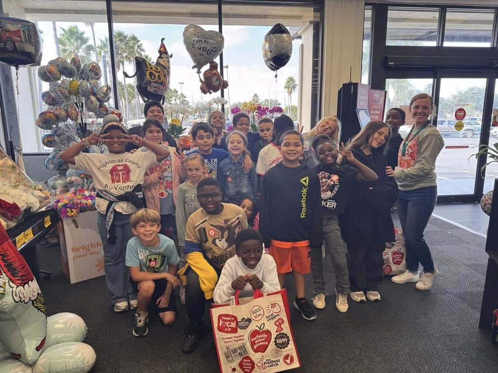 A group of approximately 15 elementary school children and two adult women pose smiling for a photo inside the front entrance of a grocery store, surrounded by displays of balloons and flowers.