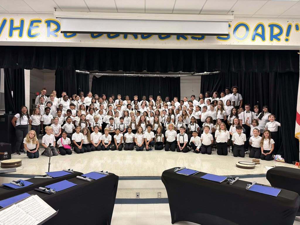 A very large group of elementary-aged students, all dressed in white shirts and black pants/skirts, are posing on a stage in front of black curtains. A banner above the stage reads, "WHERE EAGLES SOAR!"