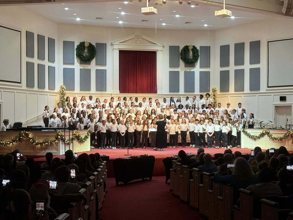 The same large group of formally dressed students is performing as a choir on a stage in a large auditorium or church, singing under a conductor while facing a seated audience.