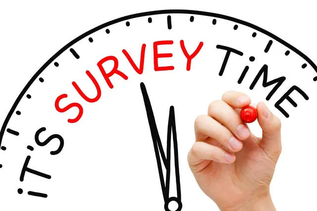 A hand holding a red marker writes the words โSurvey Timeโ in red letters across the top of a clock graphic, suggesting that it is time to participate in a survey. The black clock hands point upward toward the text.