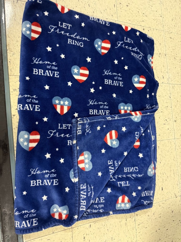 A dark blue, plush blanket is folded on the floor. It is decorated with red, white, and blue hearts, white stars, and the text "LET Freedom RING" and "Home of the BRAVE."