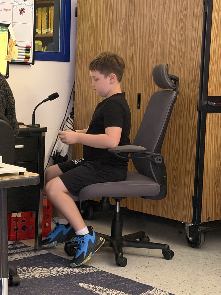 : A boy in all-black attire and blue/black sneakers sits in an office chair, looking down at a small object in his hands next to a desk microphone.
