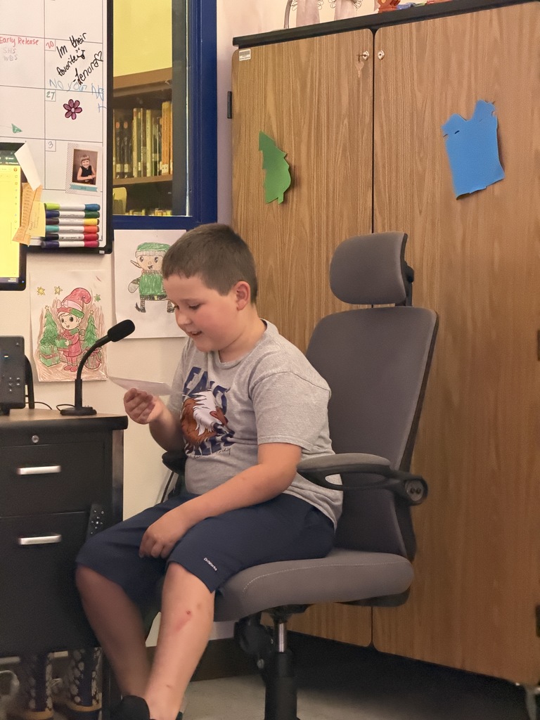 A boy in a grey t-shirt and shorts sits in an office chair, smiling while reading from a small paper into a desk microphone in a classroom.