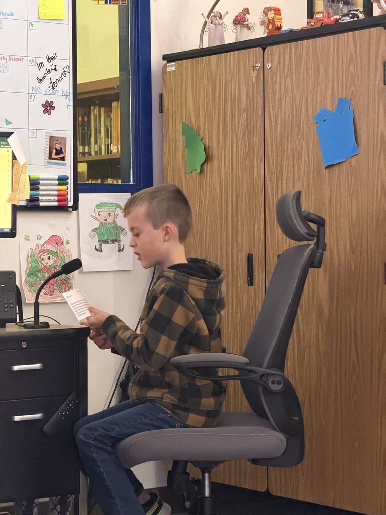 A boy in a brown and black plaid jacket and jeans sits in an office chair, focused on reading from a small paper into a desk microphone.