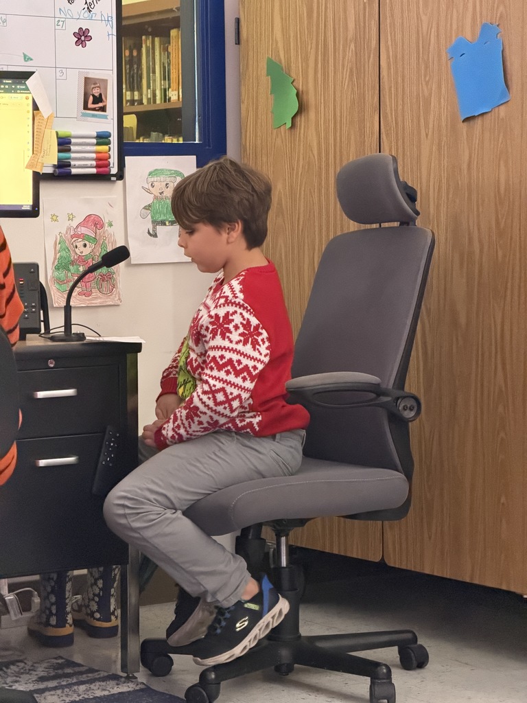 A boy in a red and white holiday sweater and grey pants sits in an office chair, looking focused while preparing to speak into a desk microphone in a classroom.