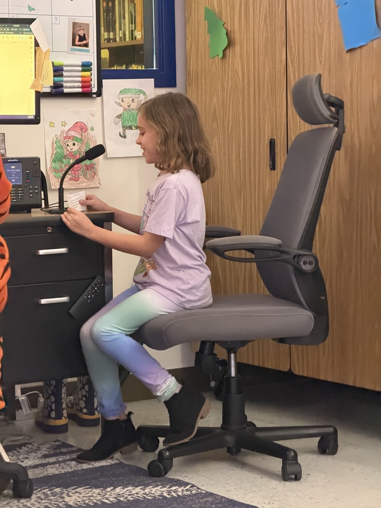 A girl in a lavender shirt and colorful leggings sits in an office chair, smiling and reading from a small paper into a desk microphone in a classroom.