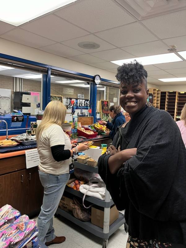 A woman with dark, styled hair and a black wrap smiles at the camera. In the background, a woman with blonde hair serves food from a breakfast buffet. A utility cart sits between them in a school-like setting.