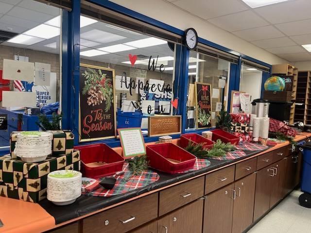 A long counter features a festive breakfast spread with "We Appreciate You" and "'Tis the Season" signs. The setup includes red serving bins, stacks of bowls, holiday greenery, and wrapped gifts. Plaid placemats decorate the surface.