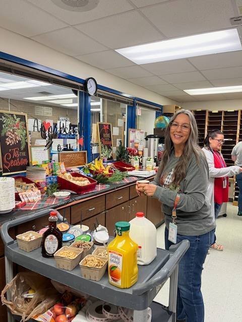 A smiling woman with long gray hair holds a cup in a lounge area. Behind her is a holiday breakfast buffet on a counter. A utility cart in the foreground holds milk, orange juice, and various toppings in small bowls.