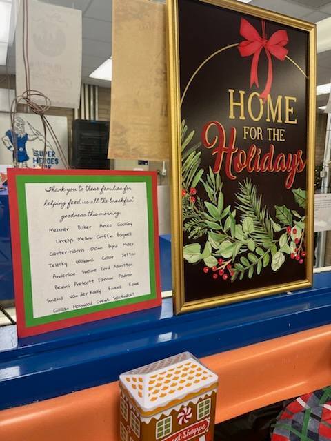 A close-up of holiday signs on a blue counter. One sign reads "Home for the Holidays," while another lists names of families to thank for providing breakfast. A gingerbread house tin is visible in the foreground.