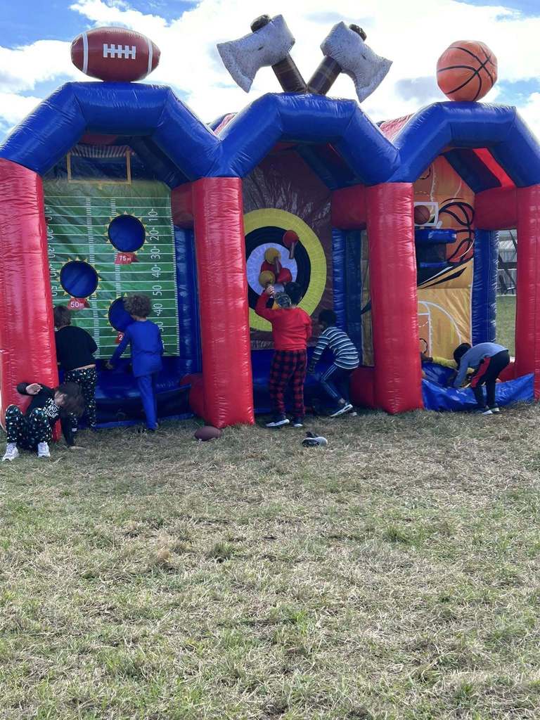 An outdoor scene featuring a large, multi-sport inflatable game structure with sections for football, axe throwing, and basketball. Several children in pajamas are actively playing at the different stations on a grassy lawn under a bright, blue sky.