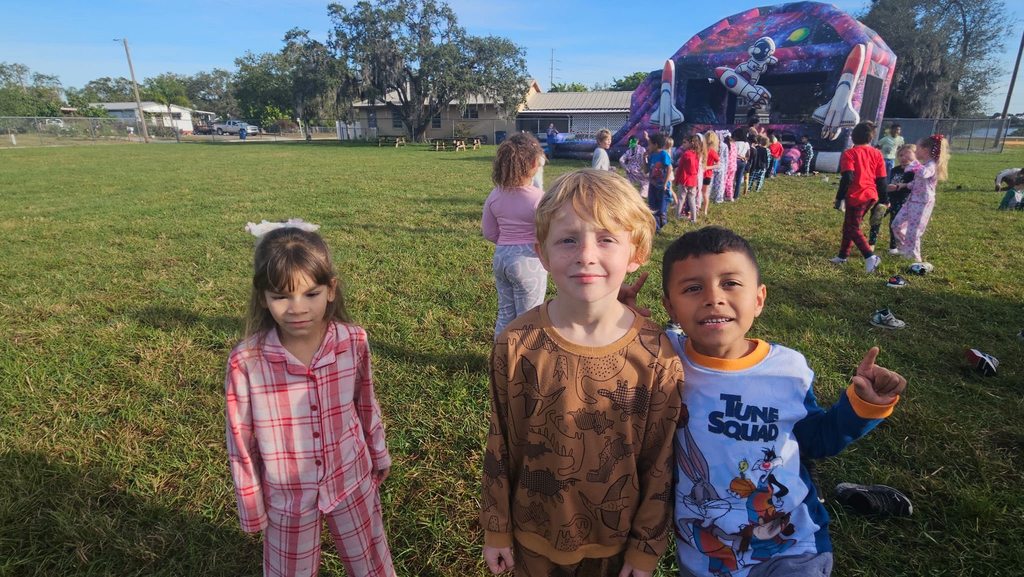 Three young children in pajamas stand in a grassy field. In the background, a large, purple space-themed inflatable slide with rockets is visible, along with other children playing. The setting is a bright, sunny day with houses and trees in the distance.