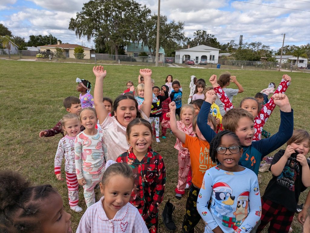A large, diverse group of young children stands on a grassy field, many wearing colorful pajamas. Several children have their arms raised high in excitement, smiling and cheering. In the background, residential houses and a large tree sit under a partly cloudy sky.