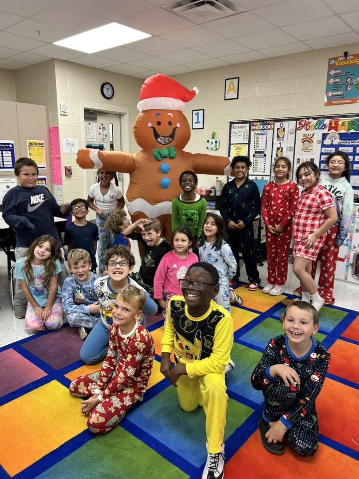 A large group of smiling children in colorful pajamas pose on a gridded rug in a classroom. Behind them stands a tall inflatable gingerbread man wearing a Santa hat. Educational posters and decorations are visible on the surrounding walls.